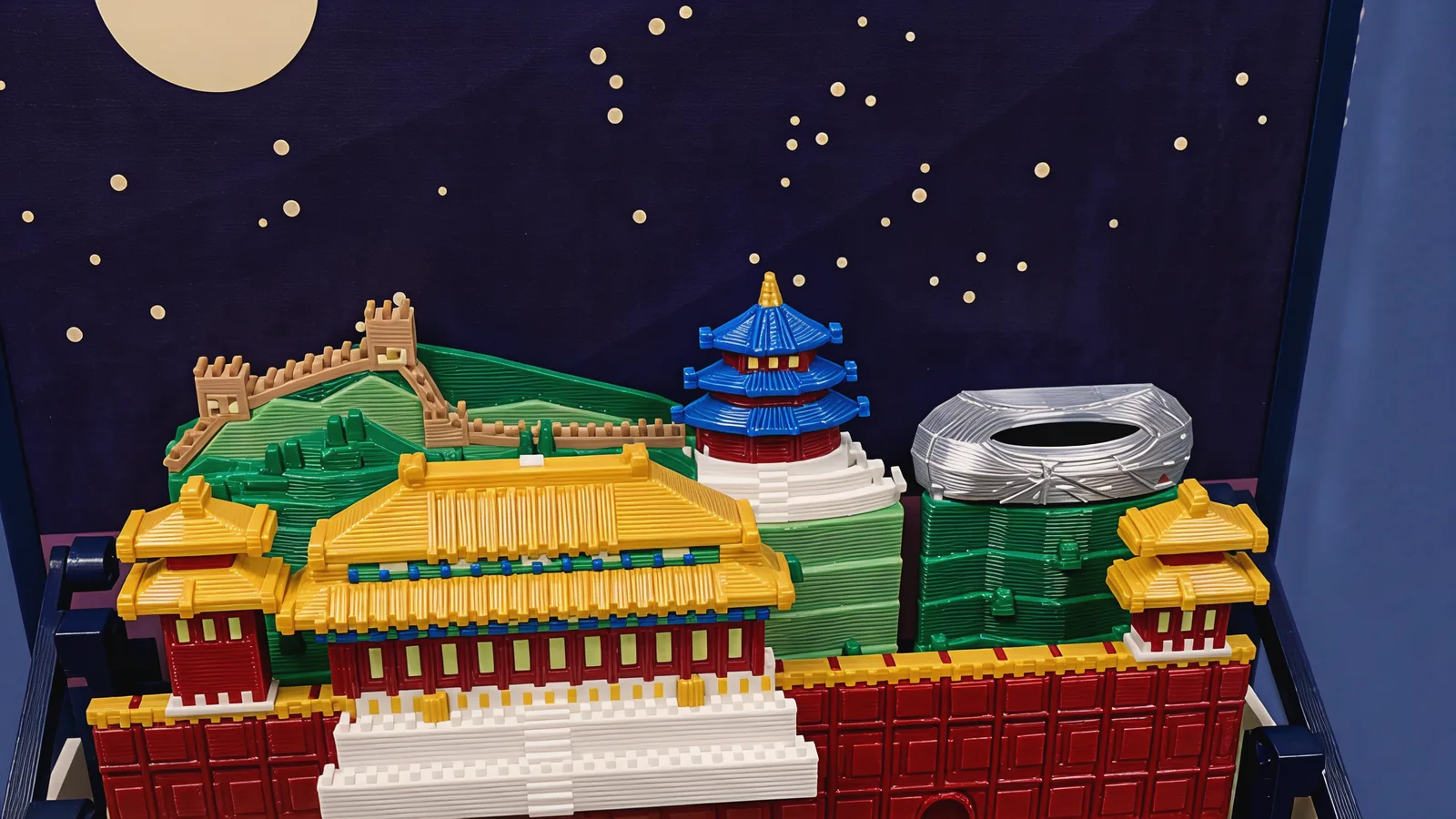 The back row against the dark indigo sky panel and cream moon: a tan crenellated wall with two watchtowers climbing green hills, a round hall with three blue roofs, a red drum and a gold tip on a white stepped base, and a silver-grey oval stadium with an open roof; in front of them the double golden roof of the palace hall and the golden roofs of the two corner towers
