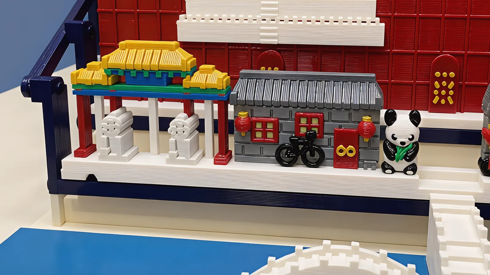 Close-up of the front row: a three-bay archway on red posts with blue and green beams and golden roofs over two pale stone lions, a grey-brick house with a dark ribbed roof, red window frames, a red door with two gold rings and a red lantern at each side, a black bicycle leaning on it, and a black-and-white panda holding a green stalk, with the red panelled palace wall and the foot of the white terrace behind
