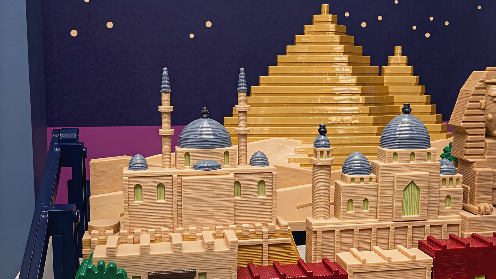 Close on the middle and back rows: a pale sandstone mosque on crenellated walls with a large grey-blue dome, small grey-blue half domes and two slim minarets with grey-blue caps, a second sandstone mosque with grey-blue domes, a round minaret and a tall pale yellow-green arched window, two golden stepped pyramids over the dunes behind them, the edge of the sphinx at the right, red roof tops at the bottom and the dark night panel with cream stars and a magenta band
