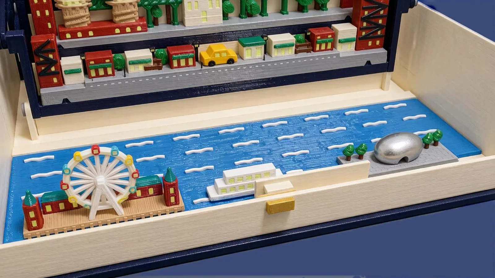 The river plate of the Chicago set in the cream tray of the open book, seen from the front left: blue water with white wave strokes; at the left a tan wooden pier with two brick-red towers under teal pyramid roofs and a long brick-red hall, and a white Ferris wheel with red, yellow and light-blue gondolas; in the middle a white tour boat with lit windows; at the right a silver bean with an arch underneath on a gray plaza between small trees; behind, the street row with brick-red and cream shops, green awnings and a yellow cab