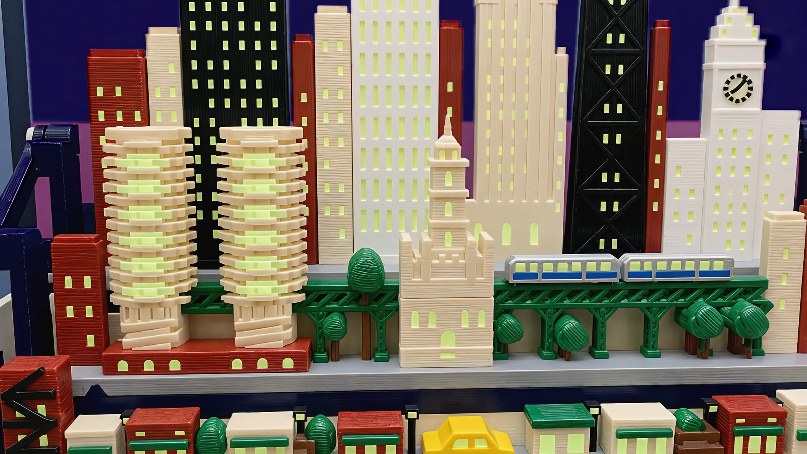 The middle row of the Chicago set: at the left two round tan towers ringed with scalloped balconies, their edges lit, on a brick-red podium with arched lit windows; a green steel elevated railway with lattice girders on arched legs, carrying two silver train cars with a blue band and lit windows; a small cream stone tower with turrets and a slim spire in front of it; round green trees; behind, black, white and cream towers with lit windows and a white clock tower; below, the street row with brick-red and cream shops under green awnings, lamp posts, trees and a yellow cab