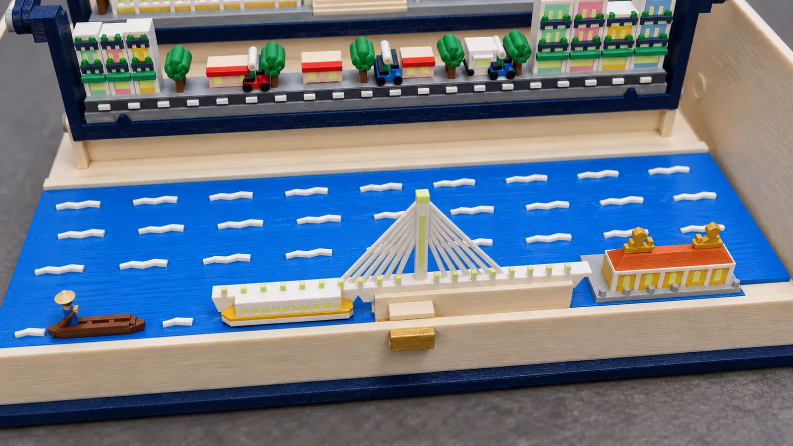 The river plate of the Ho Chi Minh City set in the cream tray of the open book, seen from above at the front: bright blue water with white wave strokes; at the left a brown wooden sampan with a rower in blue under a conical straw hat; a white water bus with a yellow band and a row of pale windows; a white bridge deck hung from fanned cables on one tall pylon with a pale yellow-green strip and tip and pale lamp caps along the deck; at the right a yellow riverside house with arched windows, a terracotta roof and two gold dragons on its ridge, on a gray quay; behind, the street row with tube houses, red awnings, trees and red and blue scooters