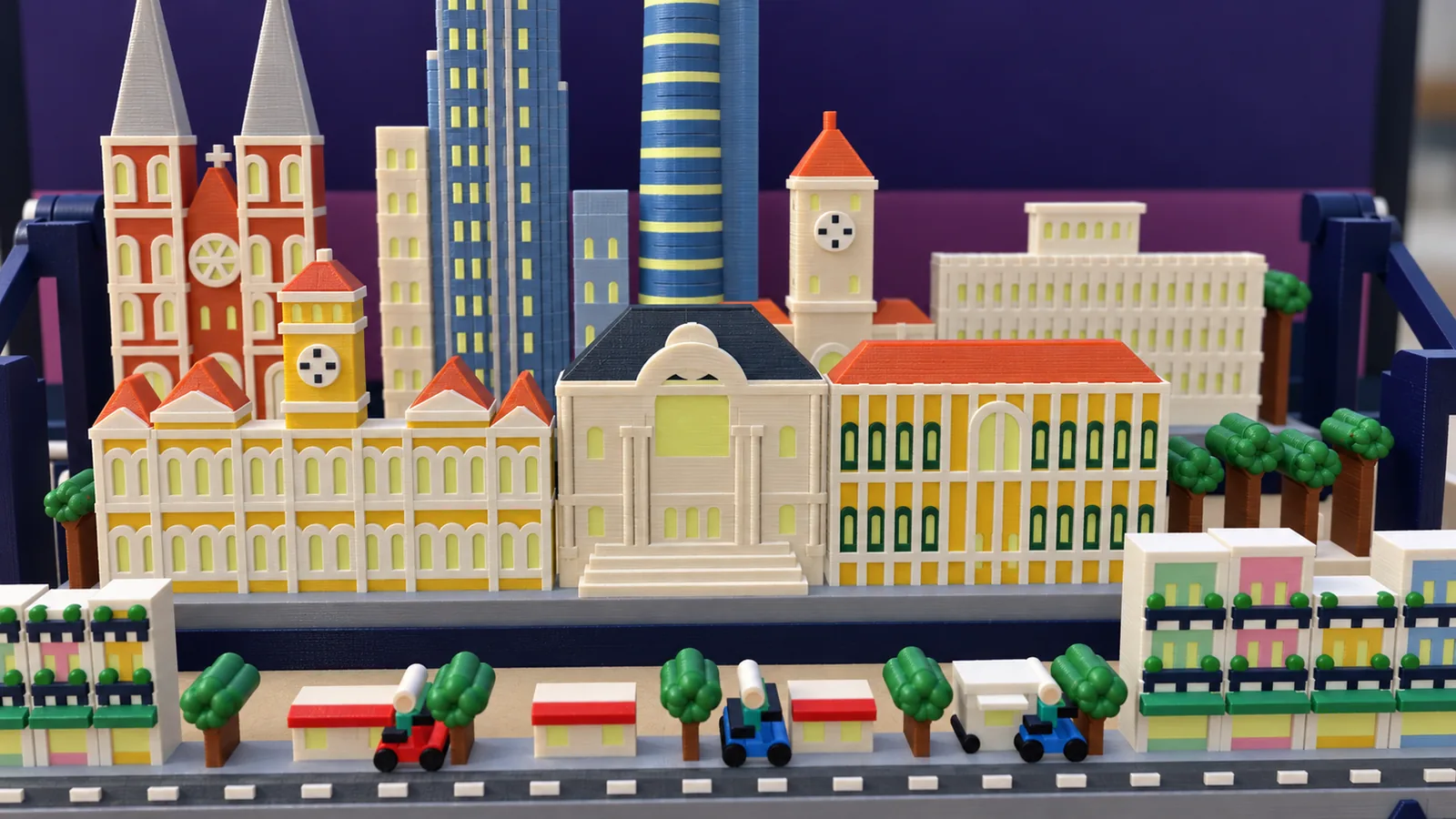 The middle and front rows of the Ho Chi Minh City set: at the left a long yellow hall with white trim, terracotta roofs and a yellow clock tower; in the middle a cream theater under a dark slate roof with a great arch, columns and a flight of steps; at the right a long yellow building with arched windows in dark green surrounds and a terracotta roof, and a park of round green trees; behind, red-brick towers under gray spires, blue towers and a cream clock tower; in front a street of narrow mint, pink, yellow and blue houses at both ends, low shops under red awnings, green trees, a white food cart and three scooters, one red and two blue, on a dark road with white dashes