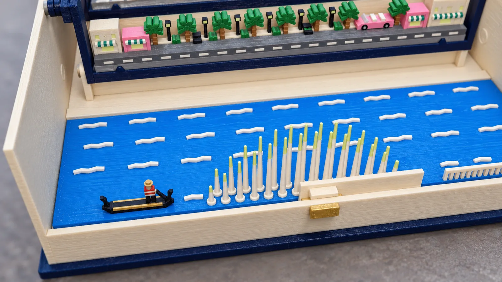 The lake plate of the Las Vegas set in the cream tray of the open book, seen from above at the front: bright blue water with white wave strokes; at the left a black gondola with gold trim and a gondolier in a red-and-white striped shirt and a gold hat; in the middle two arcs of slim white fountain jets on foam rings, with pale yellow-green tips; at the front right a low cream balustrade; behind, the boulevard row with a gray road with white dashes, cream and pink shops under striped awnings, black lamps, palms and a pink convertible