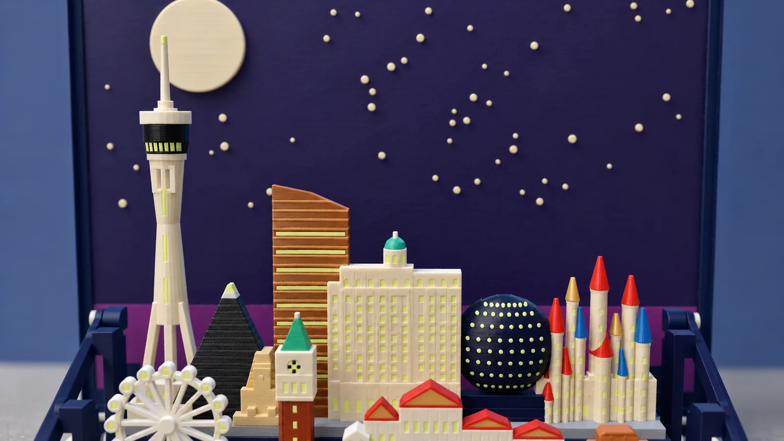 The skyline of the Las Vegas set under a large pale moon and cream stars on a dark indigo panel: at the left a tall slim cream tower on splayed legs with a dark pod ringed by lit windows and a needle reaching up to the moon; a dark pyramid with a pale tip and a sand-colored sphinx at its foot; a bronze tower banded in gold with lit bands, its roof sloping down to the right; a cream stepped building with rows of lit windows and a small teal cupola; a dark globe covered in pale glowing dots; at the right cream castle towers under red, blue and gold cones; at the bottom the top of a white spoked wheel, a brick-red bell tower with a green spire and red roofs