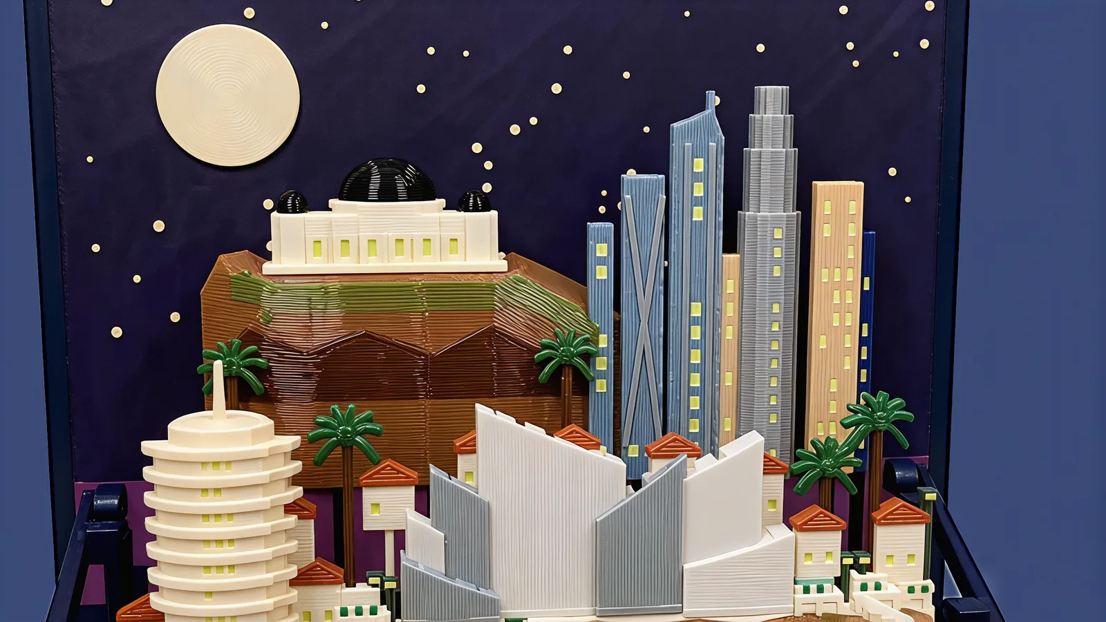 The back row against the dark indigo sky panel and cream moon: a white building with one large and two small black domes on a brown hill with an olive band, and a cluster of steel-blue, grey and sand-coloured towers with pale windows, palms in front
