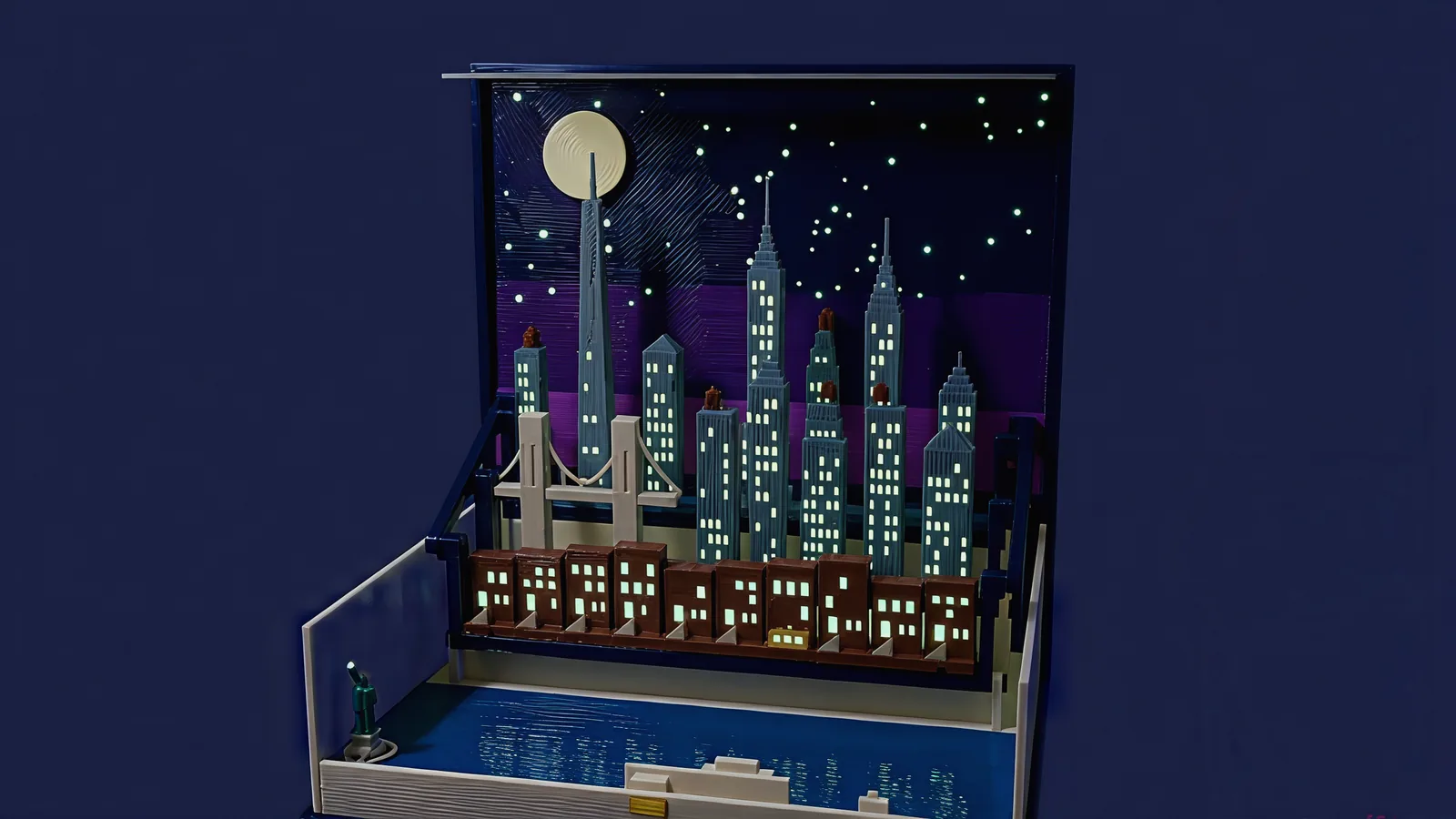 New York standing in the open navy book at night: a cream moon and glowing stars on the indigo sky panel, grey-blue towers with rows of glowing windows led by the tall spire of One World Trade Center, the pale Brooklyn Bridge on the left, a row of brown brownstones with glowing windows and a small yellow cab, and a green Statue of Liberty at the front-left corner of the dark blue harbor with a white ferry on the water