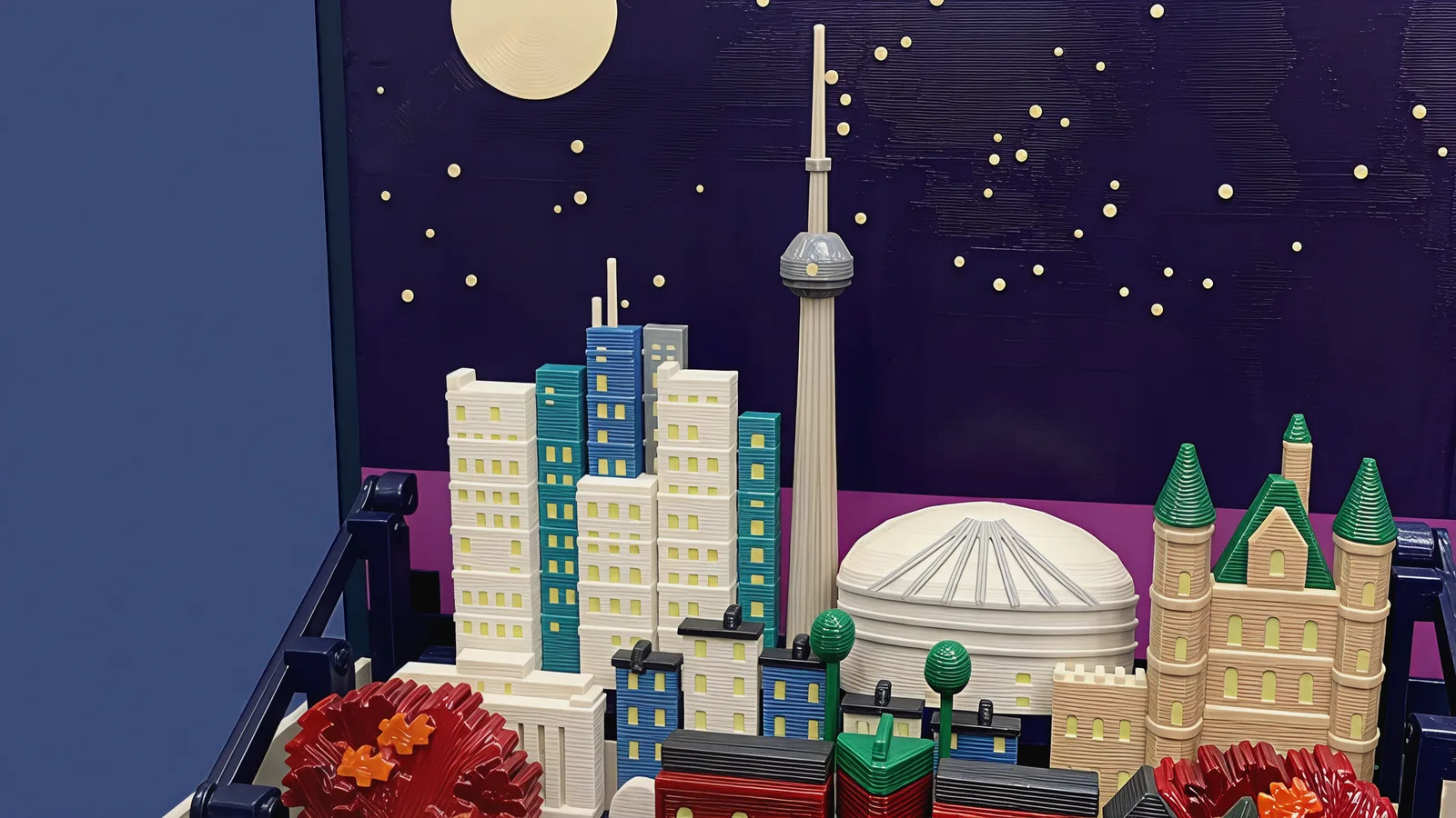 The back of the set against the dark indigo sky panel with its cream moon and stars and a magenta band along the horizon: a slim pale beige tower with a grey pod and a needle, white, blue, teal and grey towers with small pale yellow-green windows, a white ribbed dome and a sandstone castle with green cone roofs; in front, low white and blue blocks with dark caps, two round green trees and the tops of red-brick houses and red maples